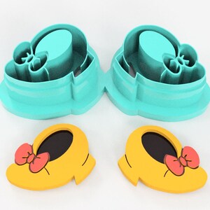 May include: Two sets of cookie cutters. The top set is teal, shaped like a shoe. The bottom set is yellow with a red bow, also shaped like a shoe. The cookie cutters are for making cookies.