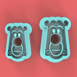 May include: Two light blue cookie cutters in the shape of a cartoon bear's face, with a round nose and eyes, and a rectangular body. The cutters are on a red background.