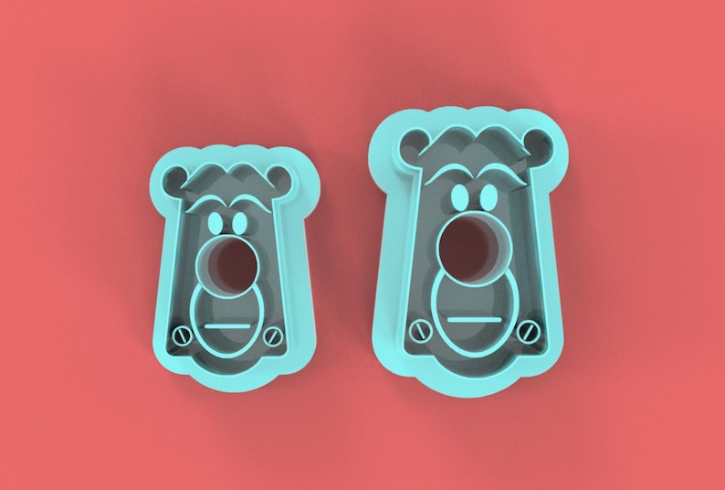 May include: Two light blue cookie cutters in the shape of a cartoon bear's face, with a round nose and eyes, and a rectangular body. The cutters are on a red background.