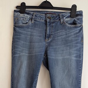 Puede incluir: Vaqueros de mezclilla azules con un diseño clásico de cinco bolsillos. Los vaqueros tienen un corte de pierna recta y cuelgan de una percha negra. La mezclilla tiene un lavado medio con decoloración y bigotes sutiles.