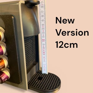 Könnte beinhalten: Dunkelgraue Espressomaschine mit schwarzer Tropfschale. Die Maschine ist 12 cm hoch. Der Text "New Version" wird auf der rechten Seite des Bildes angezeigt. Kaffeekapseln sind links sichtbar.