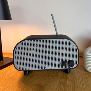 May include: A black and grey retro-style radio with a silver aerial, two knobs, and a perforated front panel. A black lamp and two decorative vases are also visible. The radio is on a wooden surface.