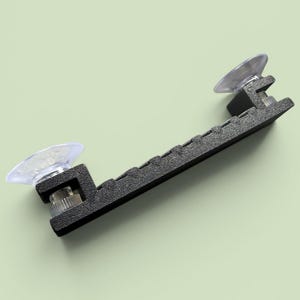 Puede incluir: Un soporte de plástico negro con dos ventosas transparentes. El soporte tiene una superficie texturizada y está diseñado para sujetar objetos en una superficie lisa. Las ventosas están unidas al soporte con un tornillo metálico.