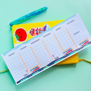 May include: A white weekly planner with a grid layout and days of the week labeled: Monday, Tuesday, Wednesday, Thursday, Friday, Saturday, Sunday. The planner has a yellow background and a blue pen on top. There are stickers on the planner, including a red tomato with a face, a colorful building, and a yellow bus.