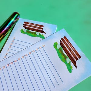 K&ouml;nnte beinhalten: Zwei Notizbl&ouml;cke mit blauen Linien und Kontrollk&auml;stchen, mit einer gr&uuml;nen Eidechsen-Grafik und dem Text "BALTIMORE THE GREATEST CITY IN AMERICA". Ein regenbogenfarbener Stift liegt auf der gr&uuml;nen Oberfl&auml;che.