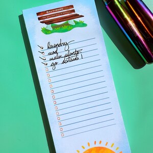 K&ouml;nnte beinhalten: Ein hellblaues Notizblock mit einer Illustration einer Parkbank und dem Text "BALTIMORE THE GREATEST CITY IN AMERICA." Das Notizblock hat eine Liste mit Kontrollk&auml;stchen und den Worten "laundry, read, water plants, go outside!" Ein regenbogenfarbener Stift liegt daneben.