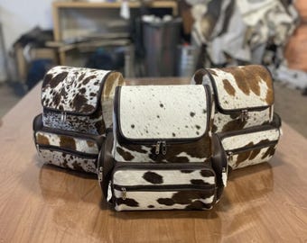 Puede incluir: Tres mochilas de piel de vaca con detalles de cuero marrón se exhiben sobre una mesa de madera. Las mochilas tienen cierre de solapa y un bolsillo frontal con cremallera. Los patrones de piel de vaca varían.