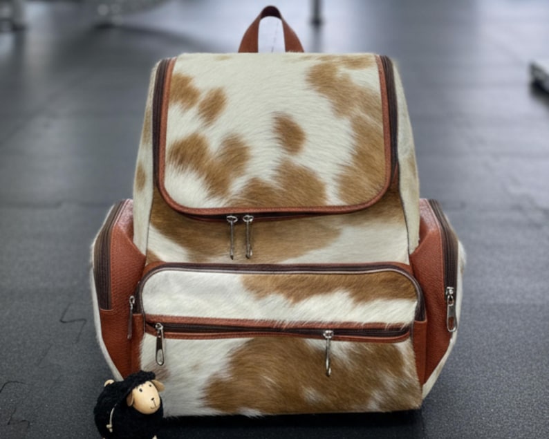 Op de afbeelding: Een rugzak van koeienhuid met een bruin-wit vlekkenpatroon. De tas heeft een klepsluiting, twee ritsvakken en bruine leren details. Een klein zwart schaapbeeldje staat voor de rugzak.