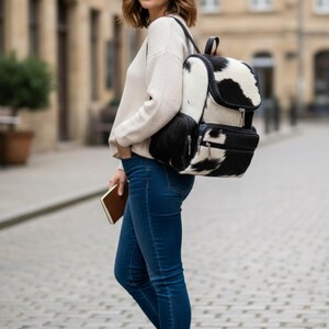 Op de afbeelding: Een zwart-witte koeienhuid rugzak met een klepsluiting en twee voorzakken. De rugzak heeft een zwart handvat en schouderbanden. De persoon op de foto draagt een cr&egrave;mekleurige trui en een blauwe jeans.