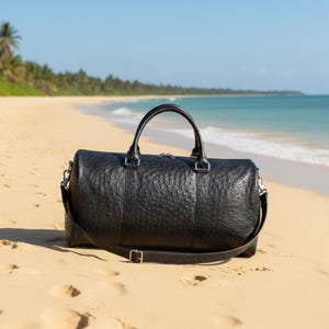 May include: A black leather duffel bag with a textured surface, a top handle, and a shoulder strap. The bag is on a sandy beach with the ocean and palm trees in the background.