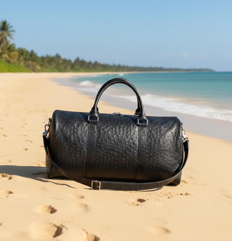 May include: A black leather duffel bag with a textured surface, a top handle, and a shoulder strap. The bag is on a sandy beach with the ocean and palm trees in the background.