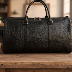 May include: Black duffel bag with a crocodile skin texture, featuring two top handles and a silver zipper closure. The bag has a rectangular shape and is sitting on a wooden surface.