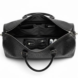 May include: Open black leather duffel bag with a textured surface. The interior reveals multiple pockets, a neatly bundled USB cable, a small case, and a pair of white earbuds. The bag has a top handle and a zipper closure.