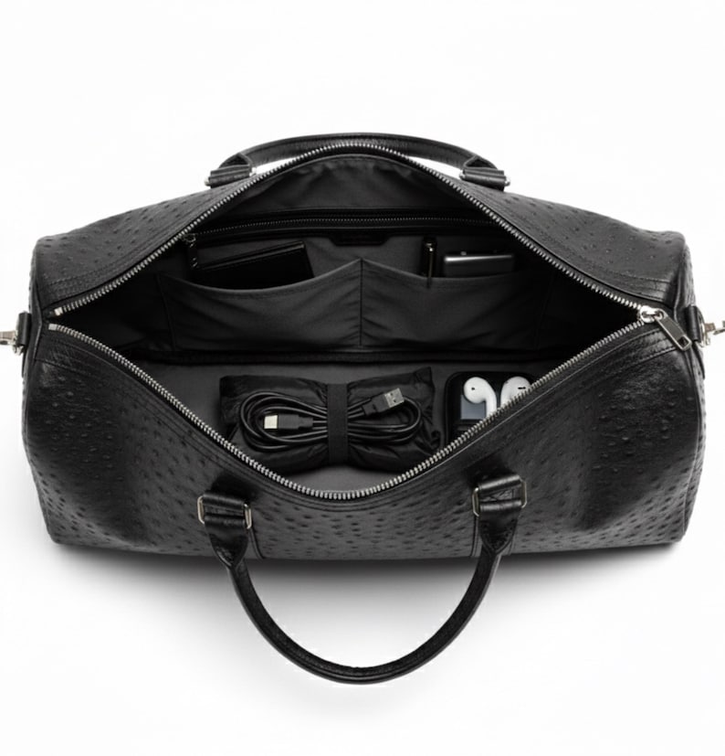May include: Open black leather duffel bag with a textured surface. The interior reveals multiple pockets, a neatly bundled USB cable, a small case, and a pair of white earbuds. The bag has a top handle and a zipper closure.