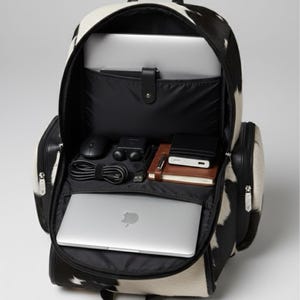 Op de afbeelding: Open zwart-witte koeienhuid rugzak met meerdere compartimenten. De rugzak bevat een laptop, muis, oplader, notitieboek en andere accessoires. De tas heeft zwarte lederen accenten en ritsen.