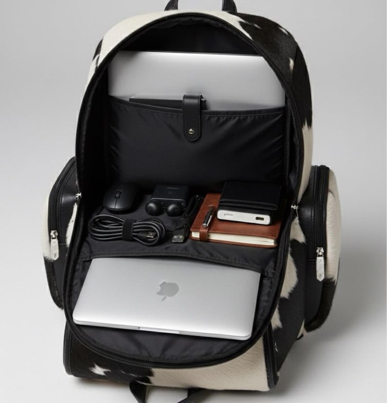 Op de afbeelding: Open zwart-witte koeienhuid rugzak met meerdere compartimenten. De rugzak bevat een laptop, muis, oplader, notitieboek en andere accessoires. De tas heeft zwarte lederen accenten en ritsen.