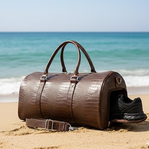 Può includere: Una borsa da viaggio marrone con una texture a pelle di coccodrillo è appoggiata su una spiaggia sabbiosa. La borsa ha un manico superiore e una tracolla staccabile. Una scarpa sportiva nera è visibile nello scomparto laterale. L'oceano è sullo sfondo.