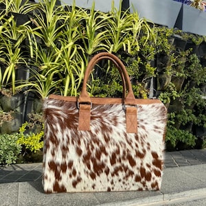 May include: A rectangular tote bag with a brown and white cowhide pattern. The bag has brown leather handles and trim, with a zipper closure. The bag is designed for carrying items and has a stylish, natural look.
