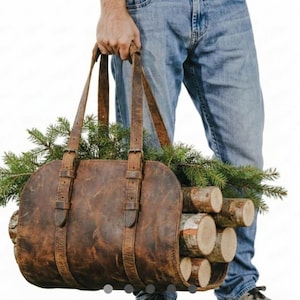 May include: A brown leather log carrier with two long straps and buckle closures. The carrier is filled with birch logs and evergreen branches. The carrier is being held by a person wearing blue jeans.