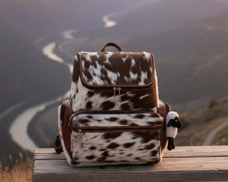 Op de afbeelding: Een rugzak van koeienhuid met bruine en witte vlekken. De tas heeft een klepsluiting, een voorvak en bruine leren details. Een klein schapenknuffel is bevestigd. De rugzak staat op een houten oppervlak met een schilderachtige achtergrond.