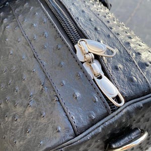 May include: Black leather duffel bag with a textured surface and silver zipper. The bag has a rounded shape and a handle on the side. The zipper is partially open, revealing the interior.