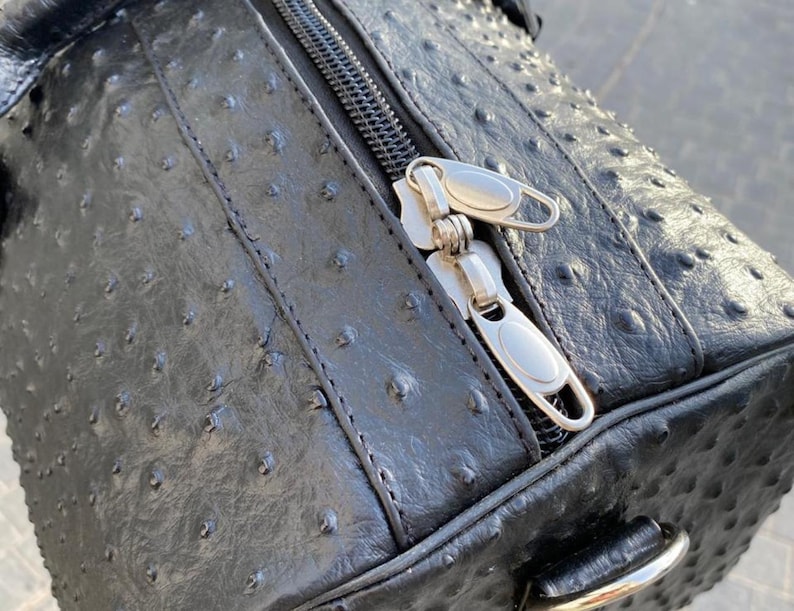 May include: Black leather duffel bag with a textured surface and silver zipper. The bag has a rounded shape and a handle on the side. The zipper is partially open, revealing the interior.