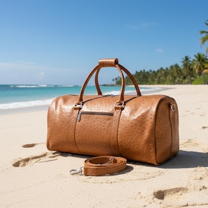 May include: A brown leather duffel bag with a textured surface, handle, and shoulder strap rests on a sandy beach. The bag has a zipper closure and metal hardware. The background features a blue sky, ocean, and palm trees.