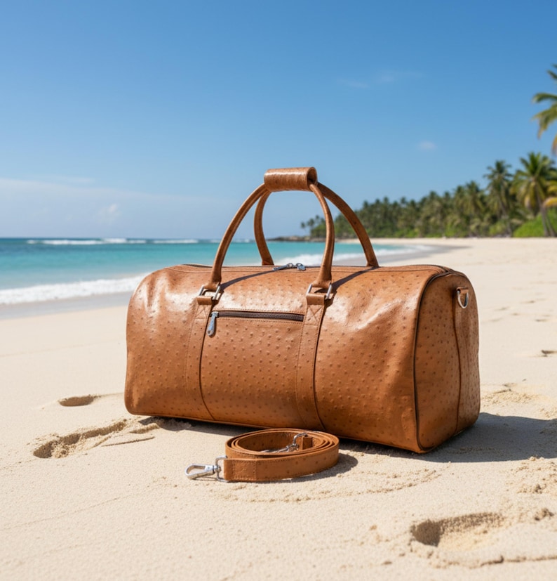 May include: A brown leather duffel bag with a textured surface, handle, and shoulder strap rests on a sandy beach. The bag has a zipper closure and metal hardware. The background features a blue sky, ocean, and palm trees.