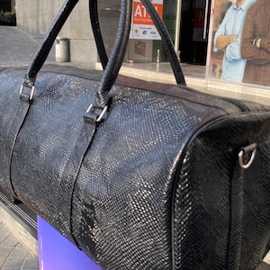 Puede incluir: Una bolsa de lona negra con un patrón texturizado similar a la piel de reptil. La bolsa tiene dos asas superiores y una fijación para correa de hombro. Los herrajes metálicos plateados acentúan el diseño de la bolsa. La bolsa está sobre una superficie morada.