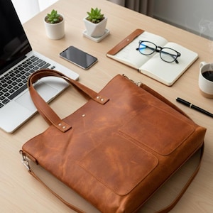 May include: A brown leather tote bag with two front pockets, a shoulder strap, and top handles sits on a wooden desk. A laptop, smartphone, notebook, glasses, pen, and small potted plants are also on the desk.
