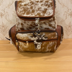 May include: A brown and white cowhide backpack with a flap closure and a front zipped pocket. The backpack has brown leather trim and a cow-shaped keyring. The bag has a top handle and adjustable straps.