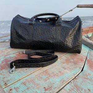 May include: A black leather duffel bag with a textured surface and a detachable shoulder strap. The bag has two handles and silver-colored metal hardware. The bag is sitting on a weathered wooden surface.
