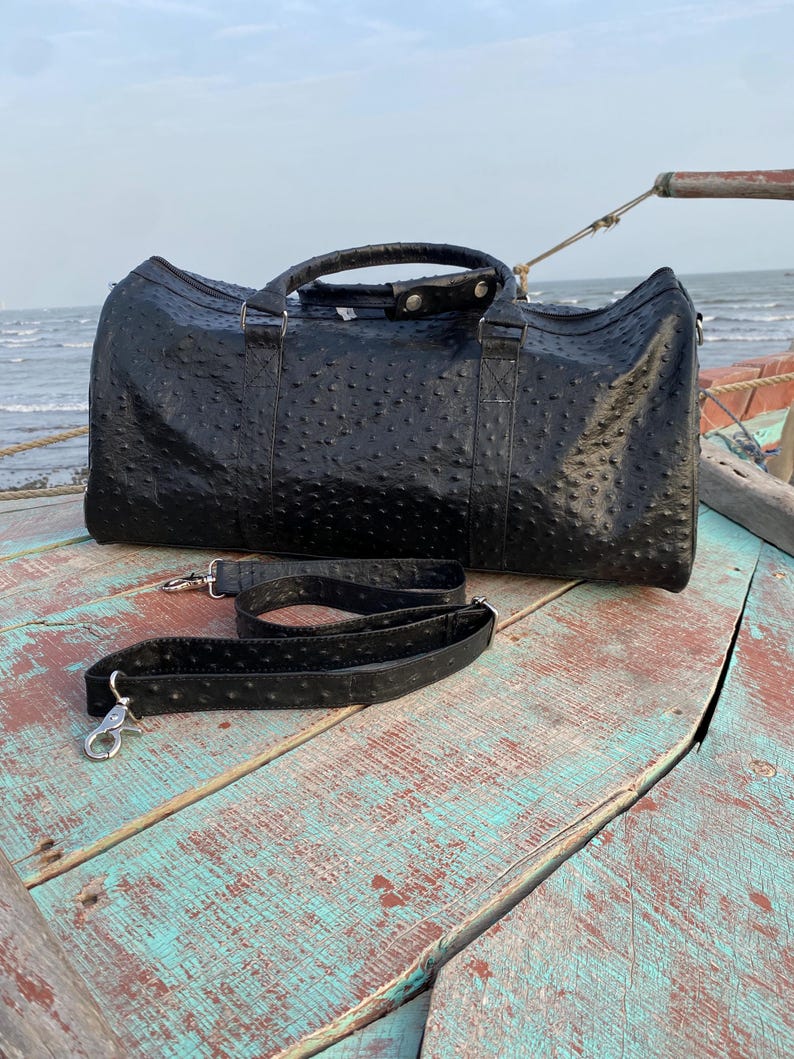May include: A black leather duffel bag with a textured surface and a detachable shoulder strap. The bag has two handles and silver-colored metal hardware. The bag is sitting on a weathered wooden surface.