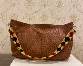 Puede incluir: Un bolso de mano de cuero tejido marrón con una correa floral colorida. El bolso es rectangular con una parte superior curvada y se apoya sobre una superficie blanca. La correa presenta flores rojas, verdes y amarillas.