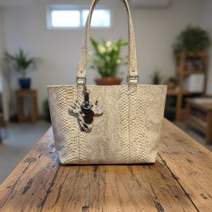 Könnte beinhalten: Cremefarbene Tragetasche mit Schlangenmuster. Die Tasche hat zwei Henkel und einen kleinen Kuhfell-Anhänger. Die Tasche steht auf einem Holztisch.
