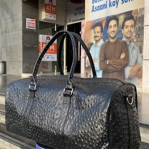 May include: Black leather duffel bag with a textured, patterned surface. The bag has two top handles and a silver-colored metal side attachment. The bag is cylindrical in shape and appears to be new.