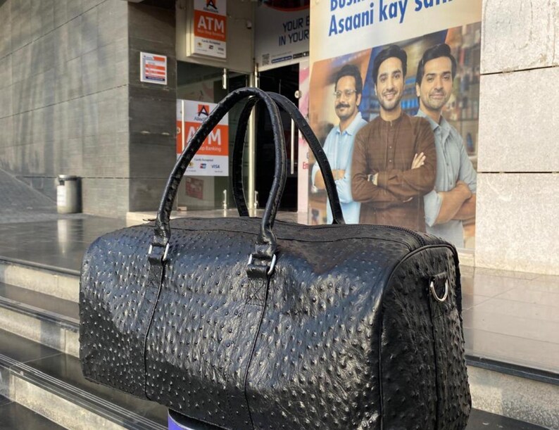 May include: Black leather duffel bag with a textured, patterned surface. The bag has two top handles and a silver-colored metal side attachment. The bag is cylindrical in shape and appears to be new.