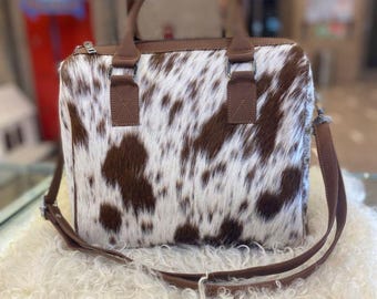 May include: A rectangular handbag with a cowhide pattern in brown and white. The bag has brown leather handles and a detachable shoulder strap. The bag is sitting on a fluffy, white surface.