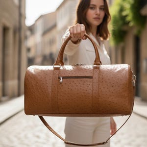 May include: A brown leather duffel bag with a textured surface and a zipper pocket. The bag has two top handles and a detachable shoulder strap. The bag is held up in front of a blurred background.