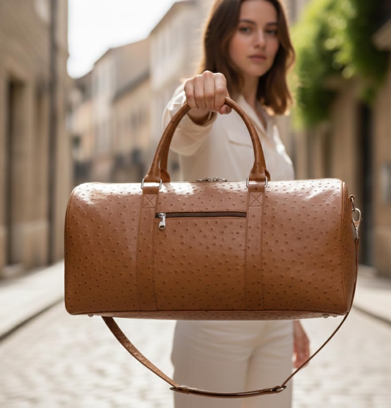 May include: A brown leather duffel bag with a textured surface and a zipper pocket. The bag has two top handles and a detachable shoulder strap. The bag is held up in front of a blurred background.