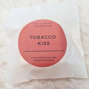 May include: A white paper package with a red circular label. The label reads "TOBACCO KISS" in white text, with "THE SENTRY COLLECTIVE" above and "luxury scented soy wax melts hand poured in Ireland" below. The package is slightly translucent.