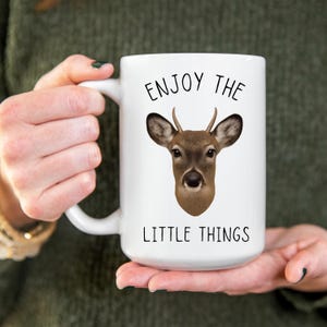 May include: White ceramic mug featuring a deer head illustration with the text "ENJOY THE LITTLE THINGS" in black. The deer has brown fur and small antlers. The mug has a large handle.