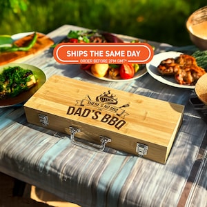 May include: A light-coloured wooden BBQ tool case with the text "THERE'S NO BBQ LIKE DAD'S BBQ" in black. The case has a silver handle and latches. The image is taken outdoors with food on plates in the background.