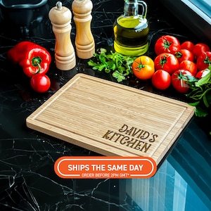 Può includere: Un tagliere rettangolare in bambù con la scritta "DAVID'S KITCHEN" incisa. La tavola è circondata da prodotti freschi, tra cui peperoni rossi, pomodori ed erbe aromatiche. Sono visibili anche due macinini per sale e pepe in legno.