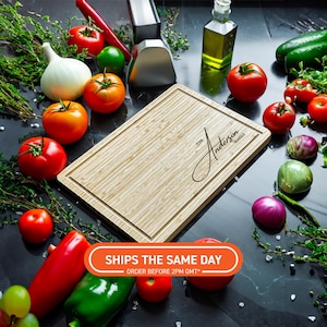 Könnte beinhalten: Ein rechteckiges Schneidebrett aus Bambus mit einer gefrästen Nut und dem Text "The Anderson Family". Das Brett ist umgeben von frischem Gemüse, Kräutern und einer Flasche Olivenöl, was auf ein kulinarisches Thema hindeutet.