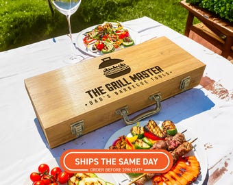 以下が含まれることがあります： 「THE GRILL MASTER - DAD'S BARBECUE TOOLS.」の文字が入った木製のバーベキューツールケースが白いテーブルに置かれています。焼き野菜や串焼きがケースの周りに並べられています。ワイングラスもあります。