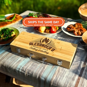 Può includere: Una valigetta per attrezzi da barbecue in bambù con la scritta "ALEXANDER'S BARBECUE TOOLS Chilling & Grilling" e un logo a fiamma. La valigetta ha una maniglia e chiusure argentate. Piatti di cibo sullo sfondo.