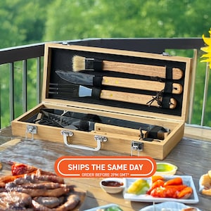 Op de afbeelding: Een bamboe barbecue gereedschapsset in een open houten kist. De set bevat een borstel, mes, vork en andere gereedschappen. De handvatten zijn houtkleurig en de kist heeft een zwart interieur. De afbeelding toont ook eten op een tafel.