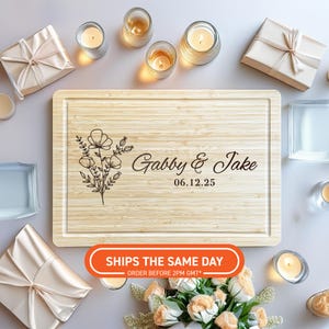 Könnte beinhalten: Ein helles Holzschneidebrett mit floralem Design und den eingravierten Namen "Gabby & Jake" und dem Datum "06.12.25". Das Brett ist von Kerzen, Geschenken und Blumen umgeben, was eine festliche Atmosphäre schafft.