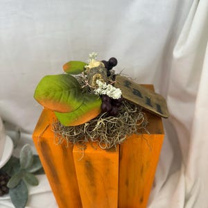 May include: A wooden pumpkin decoration painted orange with dark accents. It features a decorative arrangement of green and orange leaves, faux berries, and a small wooden tag that reads "Fall".